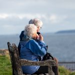 man and woman sitting on gray wooden bench viewing blue sea during daytime