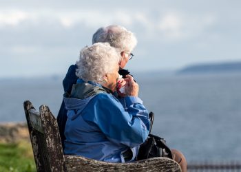 man and woman sitting on gray wooden bench viewing blue sea during daytime