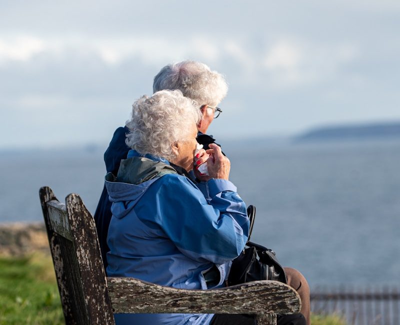 man and woman sitting on gray wooden bench viewing blue sea during daytime